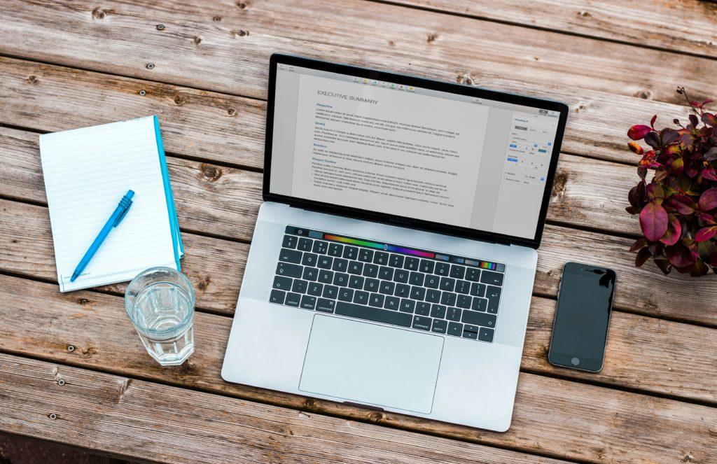 A laptop, mobile phone, notepad and glass of water on a wooden table.