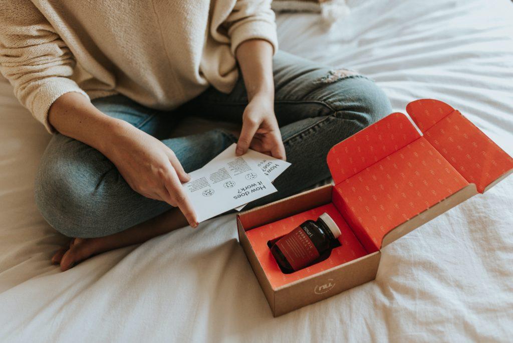 Someone sitting on a bed cross legged, holding information cards with a box with a bottle of organic supplements in front of them 